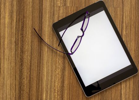 Digital tablet with blank white screen and eyeglasses on the wooden table.の写真素材