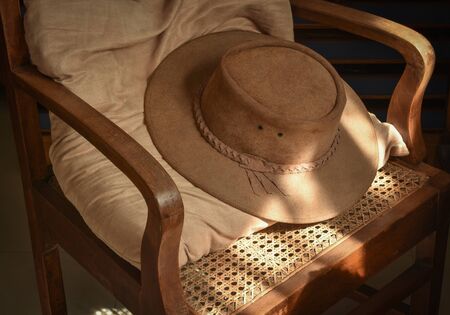 Brown suede leather cowboy hat laid on vintage wooden armchair with window light on part of it.の写真素材