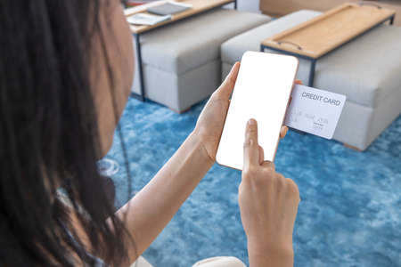 A woman shopping online on her sofa at home with a smartphone using a white credit card. And use your finger to tap the screen to verify the identity. Concept about business. Photo over shoulder shot.の写真素材