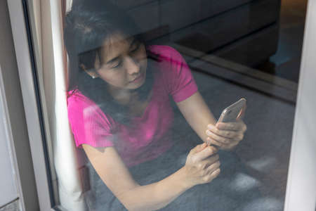 A woman sit on the floor handled a smartphone with her left hand and using her right hand's finger to touch the white screen in a house. Concept about social, technology, business. over shoulder shotの写真素材