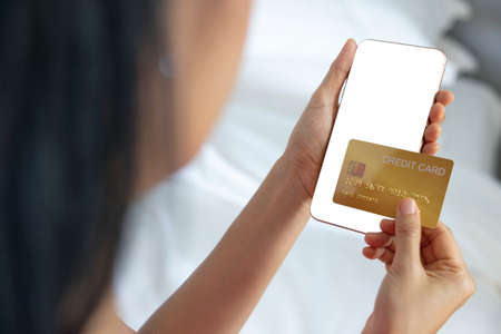 A woman shopping online on her bed at home with a smartphone using a gold credit card. And use your credit card to tap the screen to verify the identity. Concept about business. over shoulder shot.の写真素材