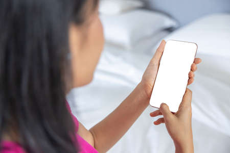 A woman lie down on a bed handled a smartphone with her left hand and using her right hand's finger to touch the white screen in a house. Concept about social, technology, business. over shoulder shotの写真素材