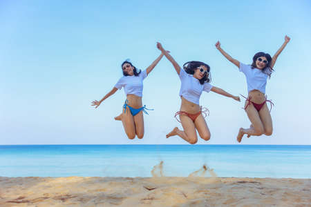 Portrait of three beautiful young asian women friends, wearing bikini and stylish sunglasses jumping on the seashore looking at camera smile. Blue sea and sky in the background.の写真素材