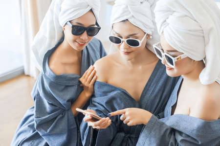 Three young woman in towel and bathrobe and sunglasses modern design sitting on bed is surfing the internet holding a phone joyfully in her hands at home.の写真素材