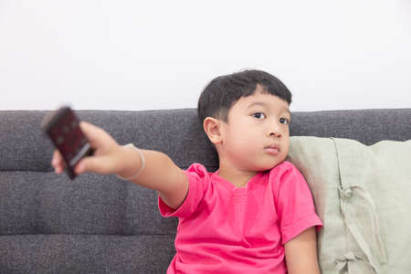 Smiling little boy watch TV and pressing the remote control on comfortable sofa in the living room. Enjoying playing game on digital smart television. Happy small kid using favorites.の写真素材