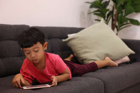 Smiling little boy looks at the touchpad lying on comfortable sofa in the living room. Enjoying playing game on digital smart phone. Happy small kid using funny applications.の写真素材