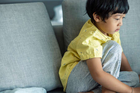 Asian boy with black hair in a yellow shirt is sitting on a tablet. Enjoying on the gray sofa in the...