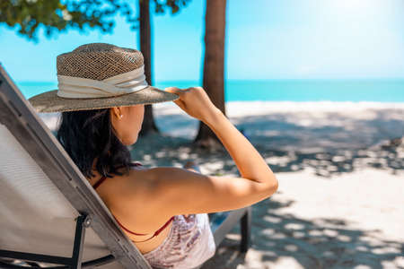 Model Asian woman wearing red swimsuit and straw hat sits in sun by the sea. Beautiful girl relaxing at infinity luxury resort. Spa, wellness.の写真素材