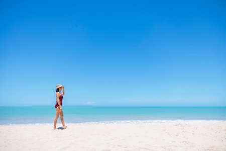 Young women in bikini and straw hat stand on tropical beach enjoying looking view of beach ocean on hot summer day. Blue sea in background. Khao Lak, Phang Nga, Thailand. Summer vacation concept.の写真素材