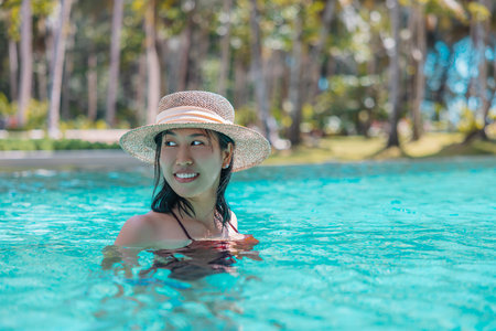 Happy asian woman in red swimsuit and a straw hat relaxing in swimming pool looking at view by the pool at Koh Mak, Phangnga, Thailand. relax and travel concept. Comfort resort summer vacation.の写真素材