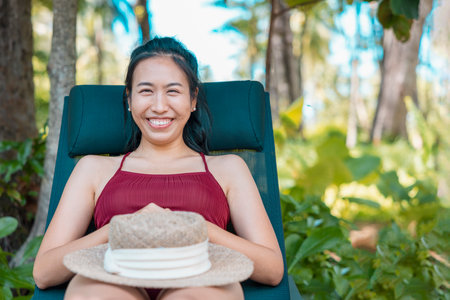 Happy asian woman looking at camera in red swimsuit and a straw hat relaxing on chair beach at Koh Mak, Phangnga, Thailand. relax and travel concept. Comfort resort summer vacation.の写真素材