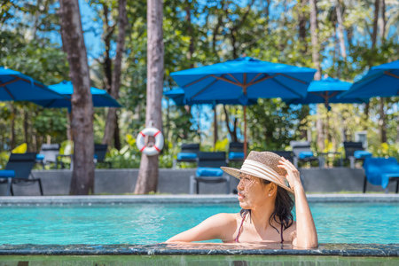 Happy asian woman in red swimsuit and a straw hat relaxing in swimming pool looking at view by the pool at Koh Mak, Phangnga, Thailand. relax and travel concept. Comfort resort summer vacation.の写真素材