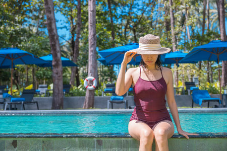 Happy asian woman in red swimsuit and a straw hat relaxing in swimming pool looking at view by the pool at Koh Mak, Phangnga, Thailand. relax and travel concept. Comfort resort summer vacation.の写真素材