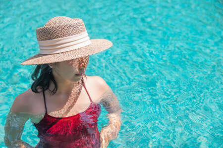 Happy asian woman in red swimsuit and a straw hat relaxing in swimming pool looking at view by the pool at Koh Mak, Phangnga, Thailand. relax and travel concept. Comfort resort summer vacation.の写真素材
