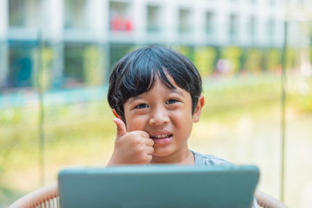 Happy Asian boy is smiling enjoying pointing thumb up sitting on the chair and play tablet. Concept of happy family or successful adoption or parenting.の写真素材
