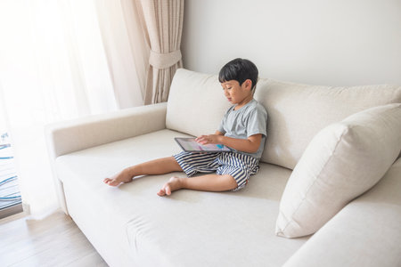 Adorable happy Asian boy wearing a gray shirt and blue-white striped shorts is having fun playing with his tablet on a cream sofa. looking at the mobile screen.の写真素材