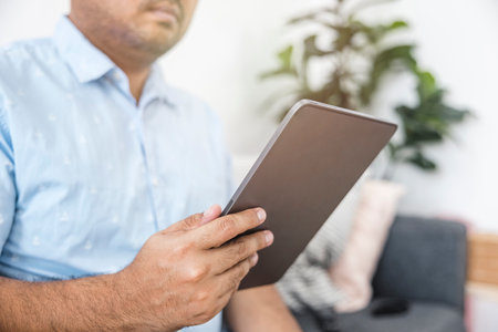 Asian man in blue shirt sitting on sofa in living room using tablet. Holiday relaxation. Online...