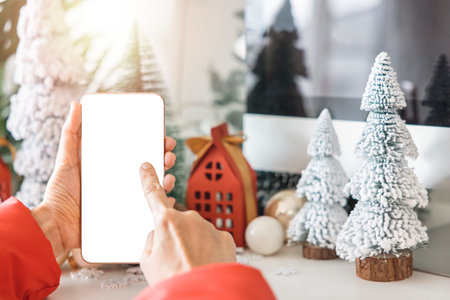 Gentle hands cradle a smartphone with a white screen mock and Fingers on the screen to shop online up by the festive scene of a beautiful Christmas tree adorned, with Christmas balls, and pine cones.の写真素材