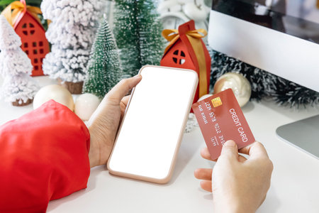 Gentle hands cradle a smartphone with a white screen mock and Hold a credit card to shop online up by the festive scene of a beautiful Christmas tree adorned, with Christmas balls, and pine cones.の写真素材