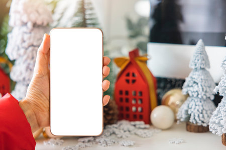 Gentle hands cradle a smartphone with a white screen mock up  by festive scene a beautiful Christmas tree adorned, with Christmas balls, pine cones, and red house gift box tied golden ribbon.の写真素材
