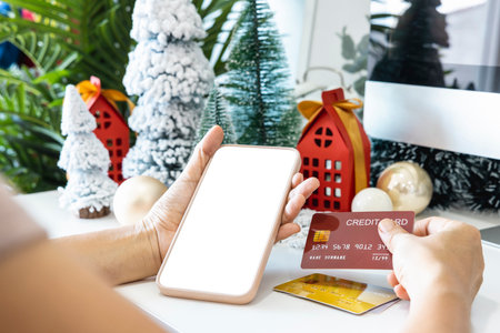 Gentle hands cradle a smartphone with a white screen mock and Hold a credit card to shop online up by the festive scene of a beautiful Christmas tree adorned, with Christmas balls, and pine cones.の写真素材