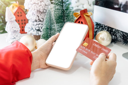 Gentle hands cradle a smartphone with a white screen mock and Hold a credit card to shop online up by the festive scene of a beautiful Christmas tree adorned, with Christmas balls, and pine cones.の写真素材