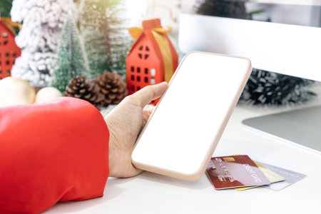 Gentle hands cradle a smartphone, empty screen by festive scene a beautiful Christmas tree adorned with credit cards, along with Christmas balls, pine cones, and red house gift box tied golden ribbon.の写真素材