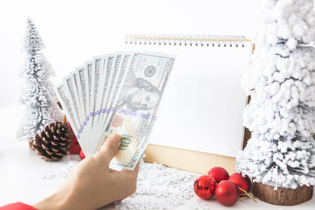 Gentle hands cradle money with a white screen mock-up and White calendar with a festive scene of a beautiful Christmas tree adorned, with Christmas balls, pine cones, and a red house gift box.の写真素材