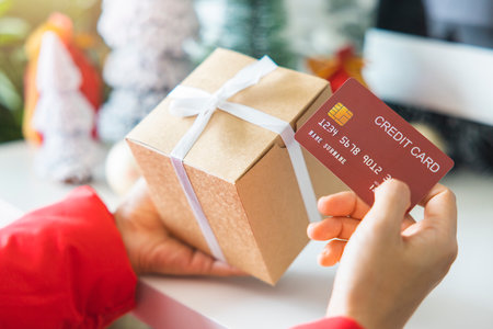 woman's hands holding a gift box and red credit cards against by the festive scene of a beautiful Christmas tree adorned, with Christmas balls, and pine cones. Buying gifts on New Year's Day.の写真素材