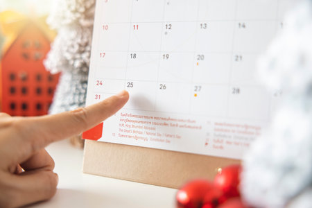A woman's fingers pointing at the calendar on December 25th, Christmas Day by the festive scene of beautiful Christmas tree adorned, with Christmas balls, pine cones. concept about Important day.の写真素材