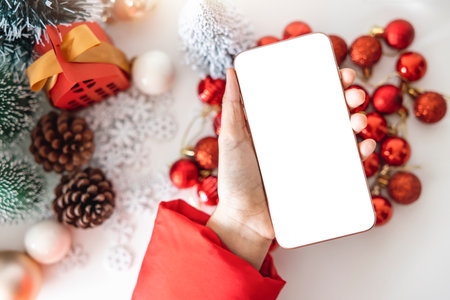 Top view Gentle hands cradle a smartphone with a white screen mock up  by festive scene a beautiful Christmas tree adorned, with Christmas balls, pine cones, and red house gift box tied golden ribbon.の写真素材