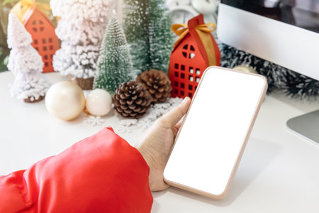 Gentle hands cradle a smartphone with a white screen mock up  by festive scene a beautiful Christmas tree adorned, with Christmas balls, pine cones, and red house gift box tied golden ribbon.の写真素材