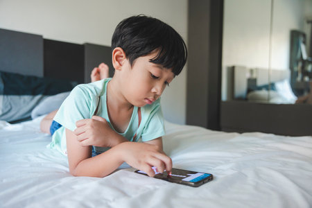 Close up portrait of Asian boy with black bangs, black eyes with a smiling face wearing a light green shirt lying on the bed in his house playing with his smart phone. Education concept.の写真素材