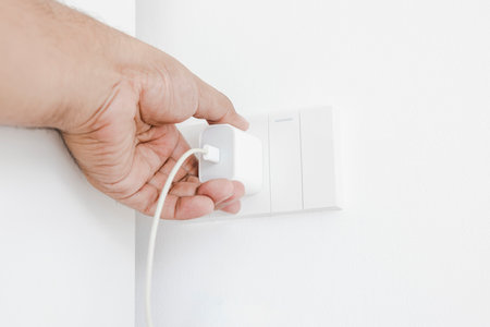 Hand plugging in an electrical appliance on white wall. Plug studio apartment power supply. Light...