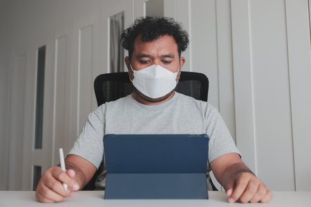 Man wearing face mask working on tablet with stylus at home, hand sanitizer on desk, concept of remote work, digital technology, and hygiene during pandemic or flu prevention.の写真素材