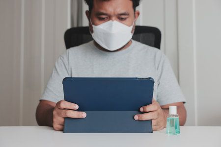 Man in face mask using tablet at home with hand sanitizer on desk, concept of remote work, online learning, digital life, hygiene, safety, and health protection during flu or pandemic.の写真素材