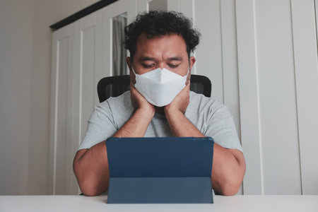 Man wearing protective face mask, sitting at desk with tablet, feeling stressed or anxious while working from home during pandemic or quarantine.の写真素材