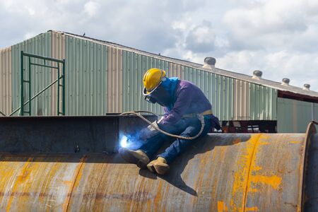The welder is welding a large steel pipe with the process Flux Cored Arc Welding (FCAW) and dressed properly with personal protective equipment(PPE) for safety, at industrial factory.の写真素材