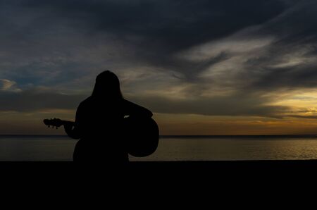 Silhouette of a woman sitting playing the guitar at the seaside In the sunset.の写真素材