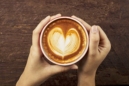 Female hands holding a cup of coffee with foam over wooden table, top viewの写真素材