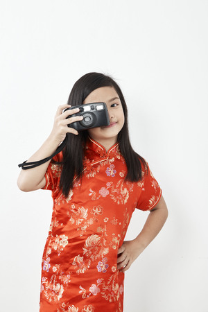 Cute little girl 8 years in Chinese dress traditional cheongsam New year 2019  takes picture with vintage retro photo camera. Doing snapshot or taking picture in red dress.の写真素材