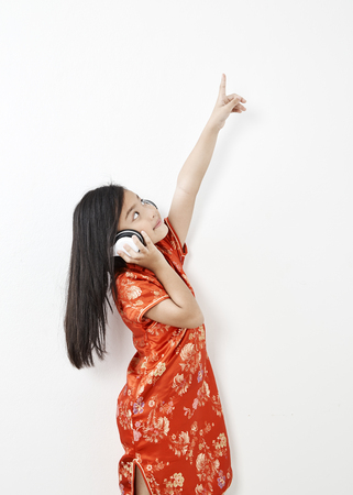 Girl child in chinese dress traditional cheongsam New year 2019 with headphones listening to music on white wall background, New Year's holiday conceptの写真素材