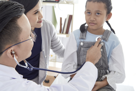 The doctor listens to the lungs of a sick boy in a stethoscope. He came with his mother to the doctor. The girl is ill with a cold. They are in the light office of the doctor.の写真素材