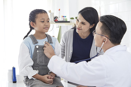 The doctor listens to the lungs of a sick boy in a stethoscope. He came with his mother to the doctor. The girl is ill with a cold. They are in the light office of the doctor.の写真素材
