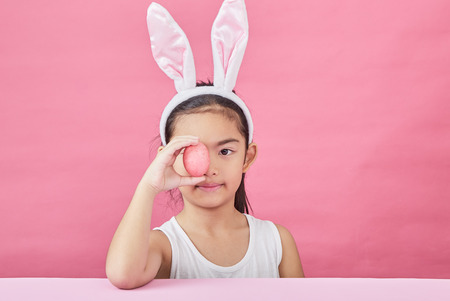 Studio shot of a happy Little girl wearing bunny ears and holding up a colorful Easter egg on a pink backgroundの写真素材