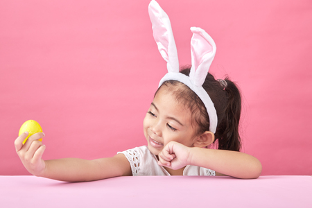 Happy asian little child girl  wearing bunny ears on Easter day. Girl holding painted eggs, pink minimal backgroundの写真素材