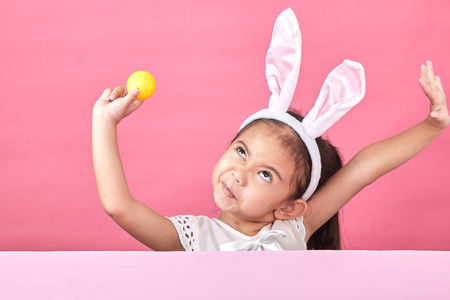 Happy asian little child girl  wearing bunny ears on Easter day. Girl holding painted eggs, pink minimal backgroundの写真素材