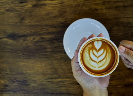 Female hands holding a cup of coffee with foam over wooden tableの写真素材