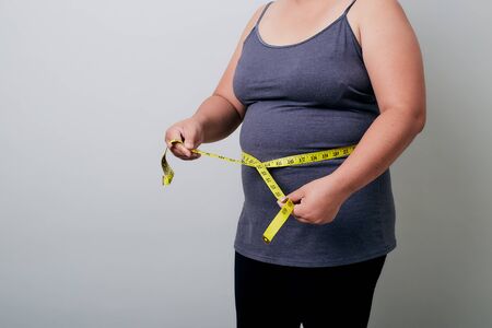 Overweight Asian woman measuring waist before weight loss in studio shot on bright gray background, health problems and overweight, diet conceptの写真素材