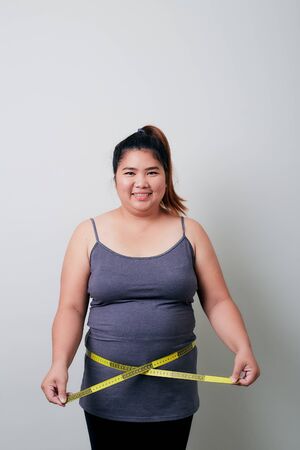 Overweight Asian woman measuring waist before weight loss in studio shot on bright gray background, health problems and overweight, diet conceptの写真素材